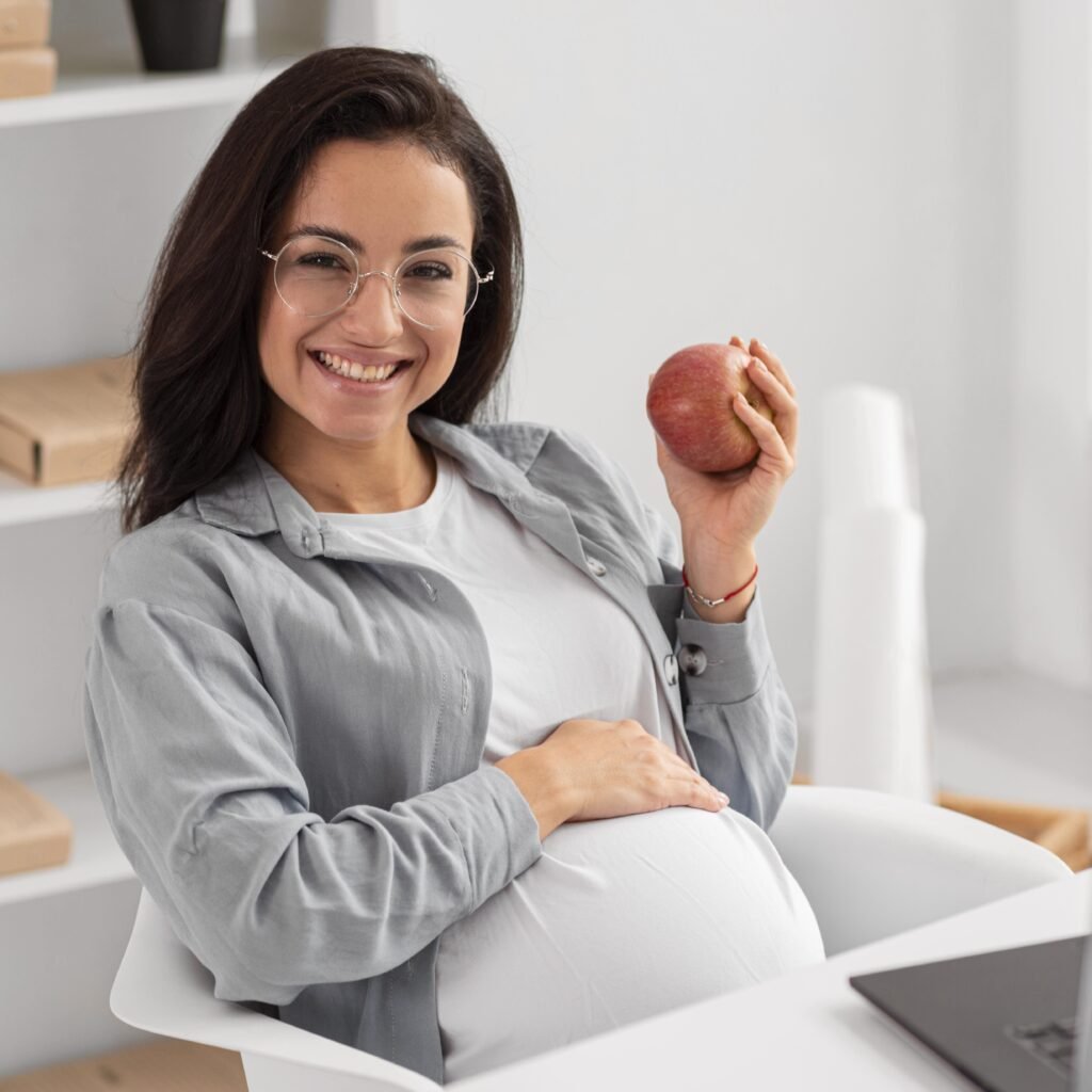 Smiling pregnant woman at home holding an apple and touching her belly, representing healthy habits and dental care during pregnancy